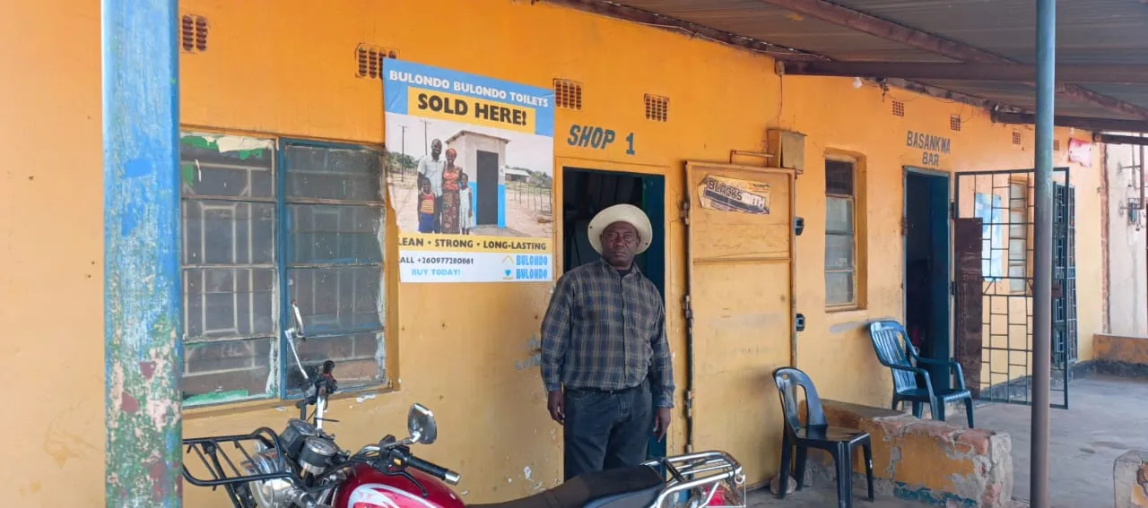 An individual stands in front of a storefront bearing a poster offering Bulondo Bulondo Latrines.