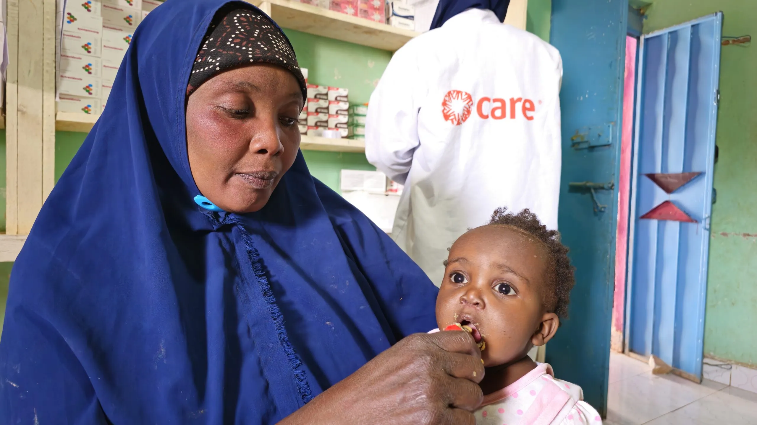 Aisha, wearing a blue hijab, feeds her 1-year-old daughter, Shukri, inside a CARE-supported health center.