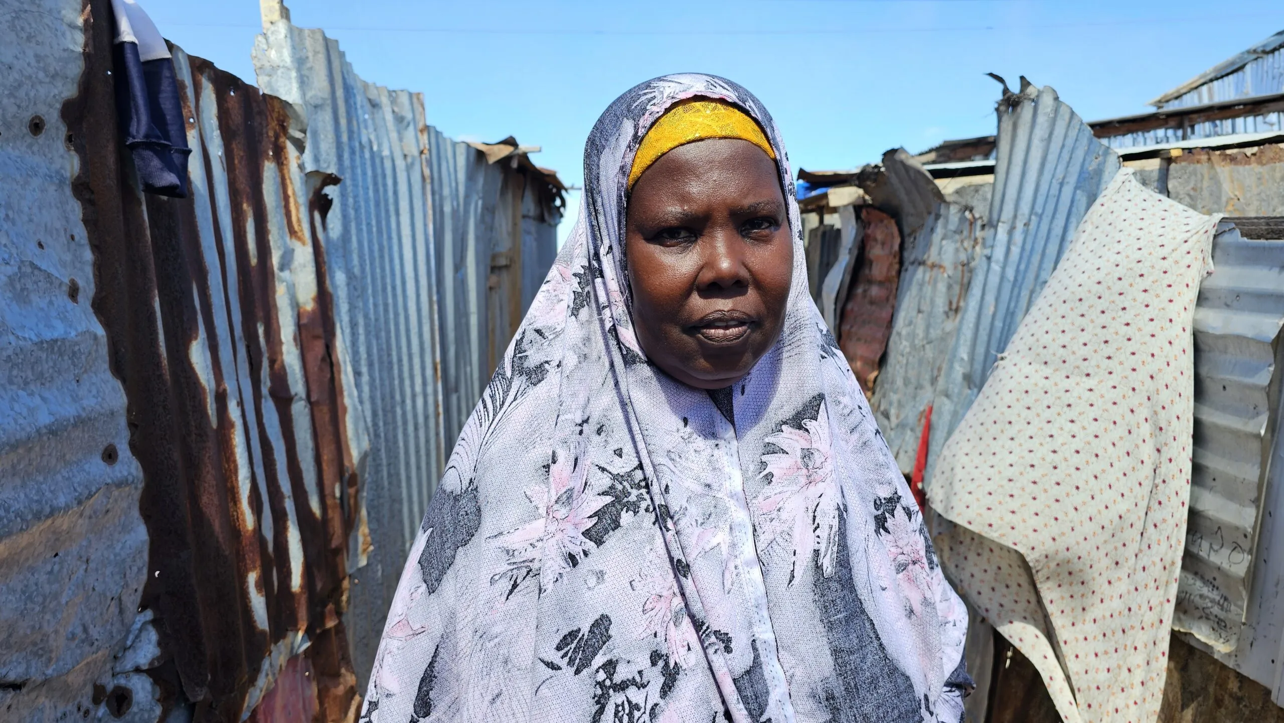 Fadumo stands between corrugated metal shelters, wearing a patterned white and lavender hijab with a yellow head covering.