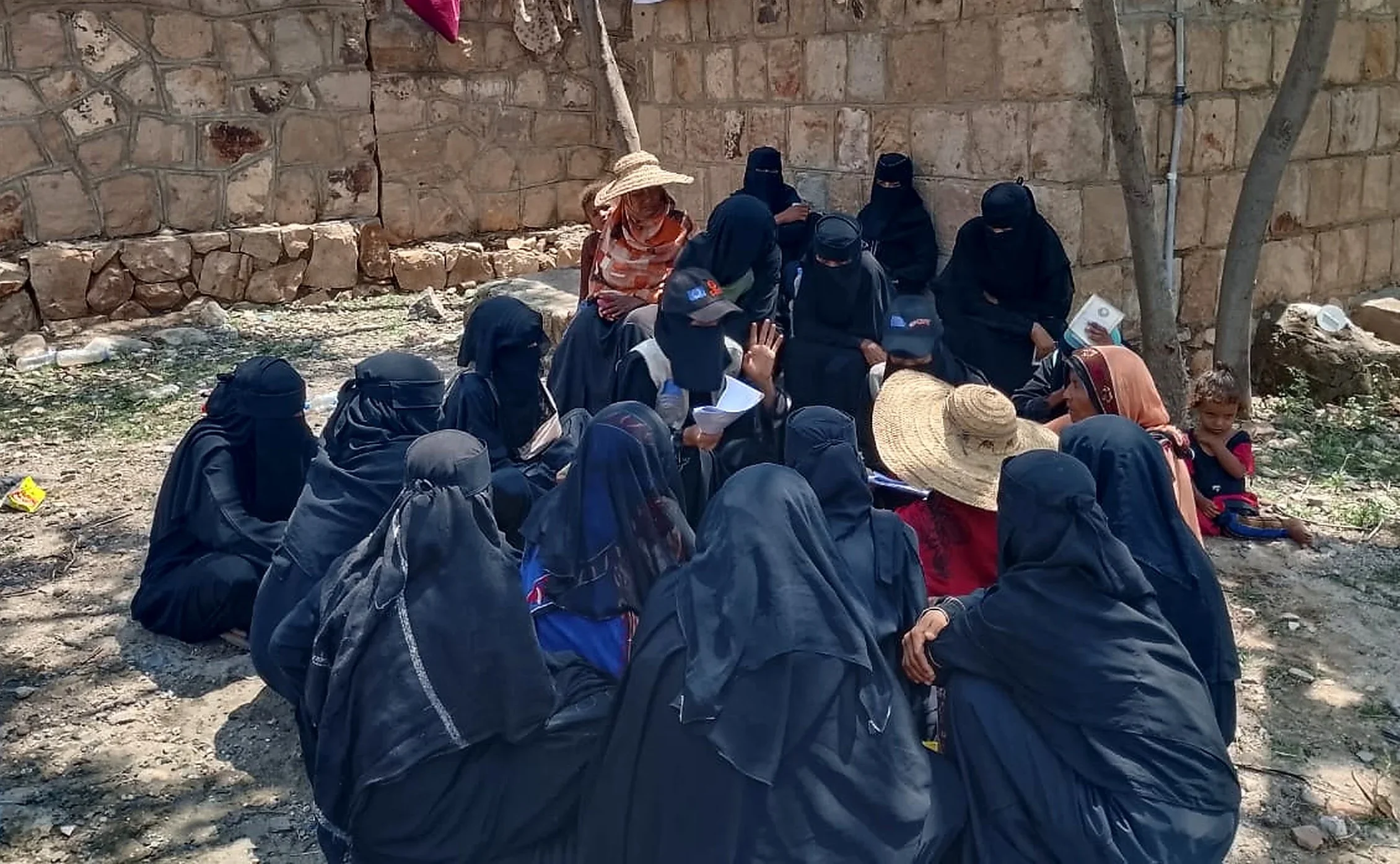 A large group of displaced women in black abayas sit in a circle outdoors at a site in Taiz governorate, Yemen, meeting with a community outreach team to discuss their needs following the September 2026 conflict escalation.