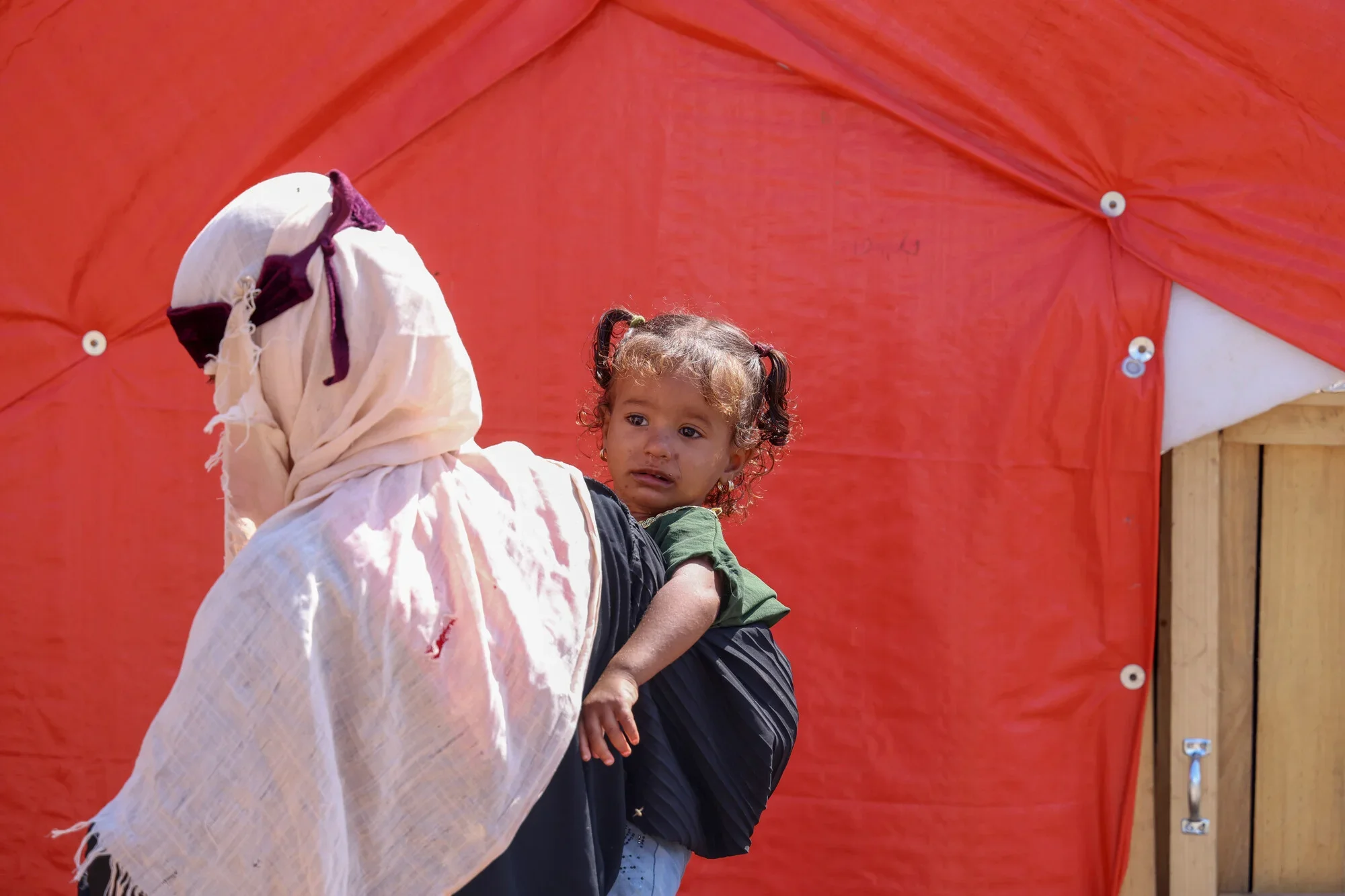 A woman in a white headscarf carries a young girl in a green top on her hip in front of a red tent at a displacement site in Taiz governorate, Yemen, as families continue to arrive following the escalation of conflict in September 2026.