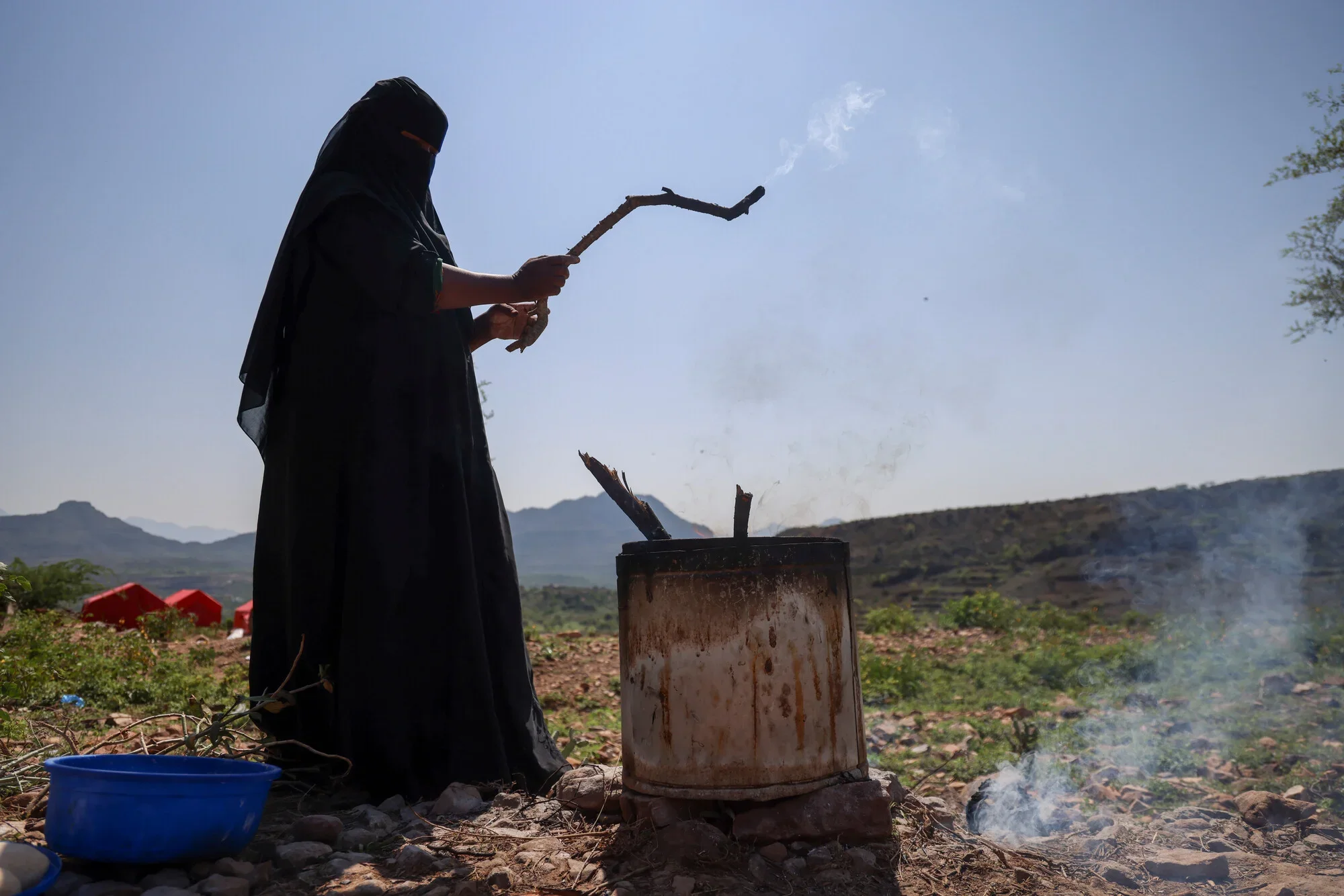 A woman in a black abaya stands outdoors stoking a fire in a makeshift metal stove using a burning stick, with smoke rising against a backdrop of mountains and red displacement tents in Taiz governorate, Yemen.