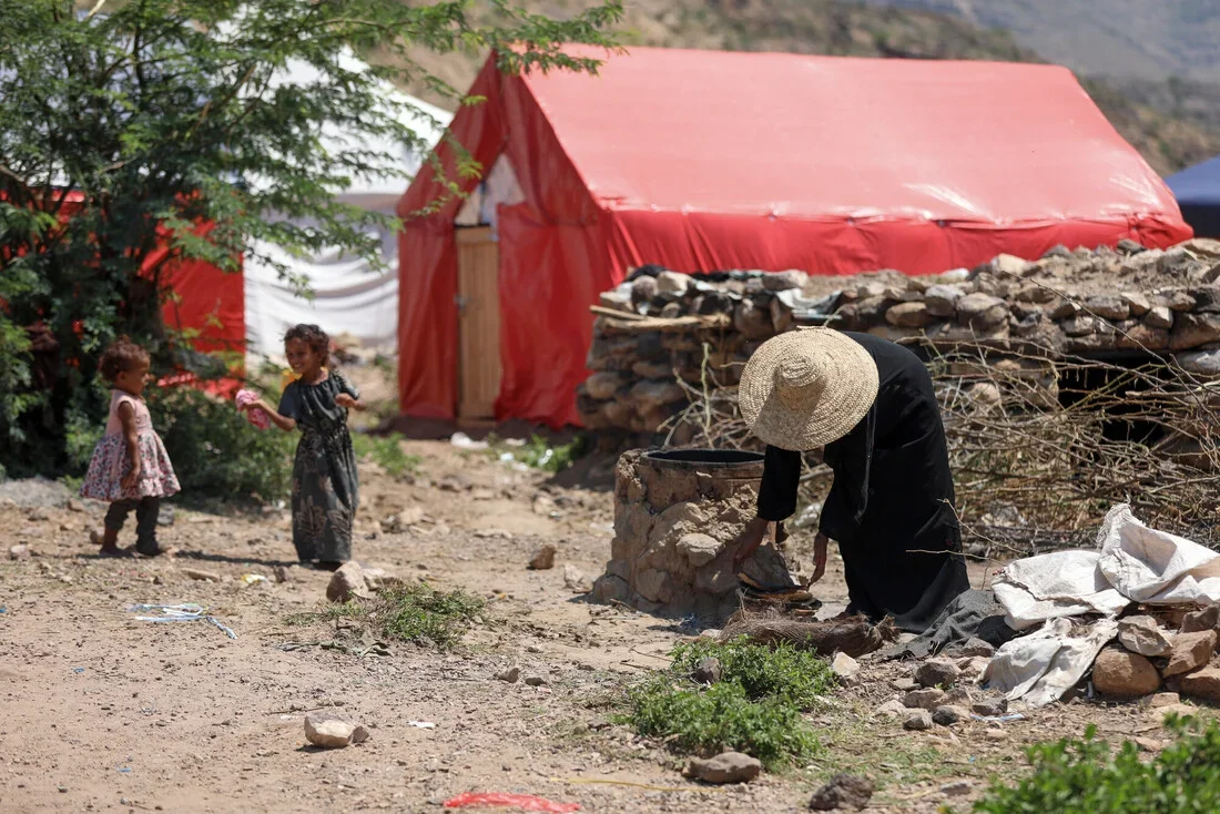 A woman in a black abaya and wide straw hat crouches over a makeshift stone stove at a displacement site, while two young girls stand nearby against a backdrop of red and white tents in Taiz governorate, Yemen.