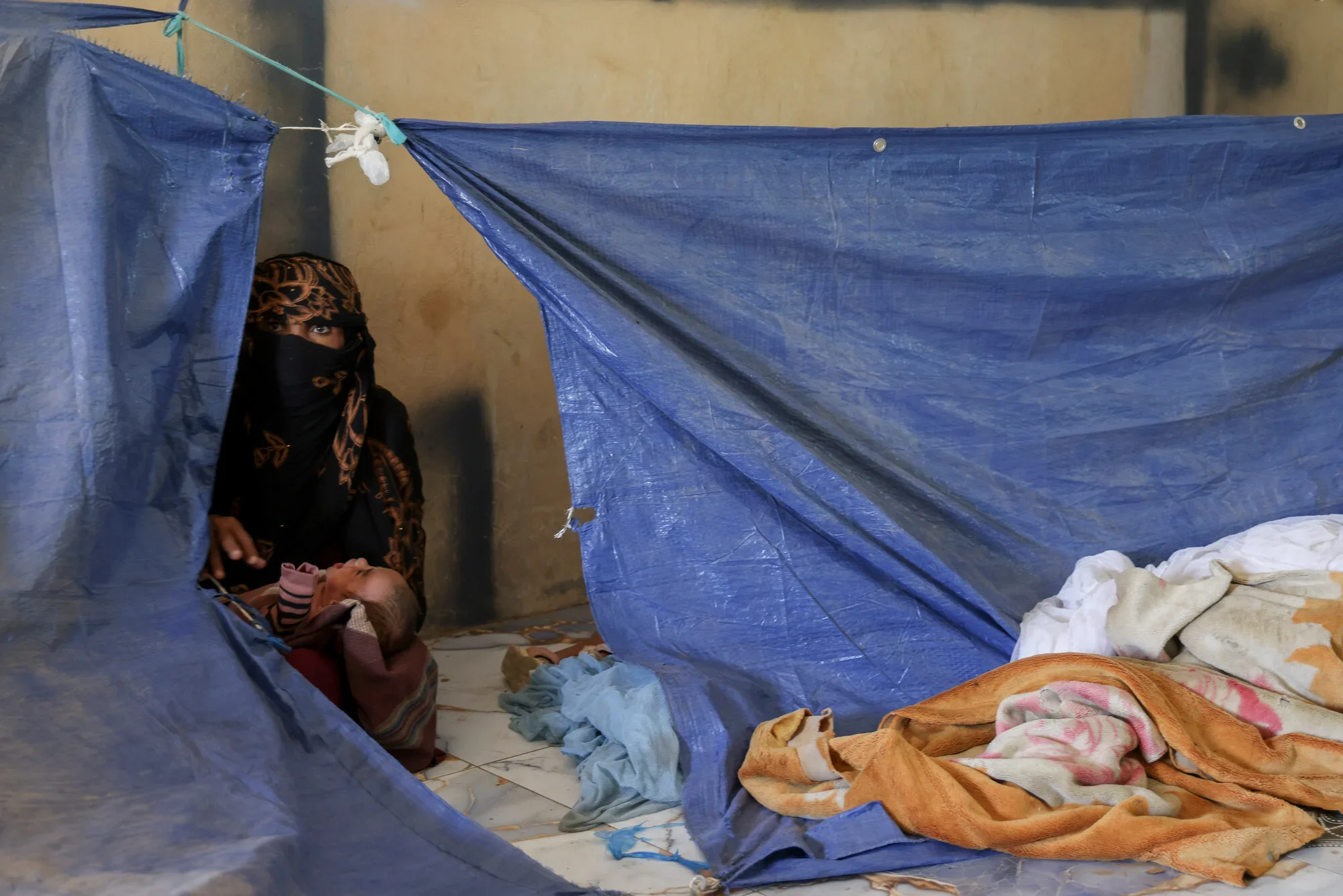 A woman in a black niqab and patterned headscarf sits inside a makeshift blue tarpaulin shelter holding a newborn infant at a displacement site in Yemen.